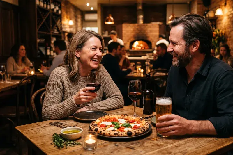 Vinello Pizzeria — a couple laughing over wood-fired pizza, red wine and a cold beer in a warm, candlelit pizzeria